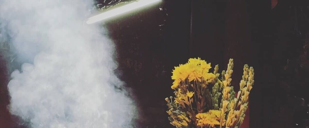 A person lighting candles at a table with a vase of flowers and incense smoke.