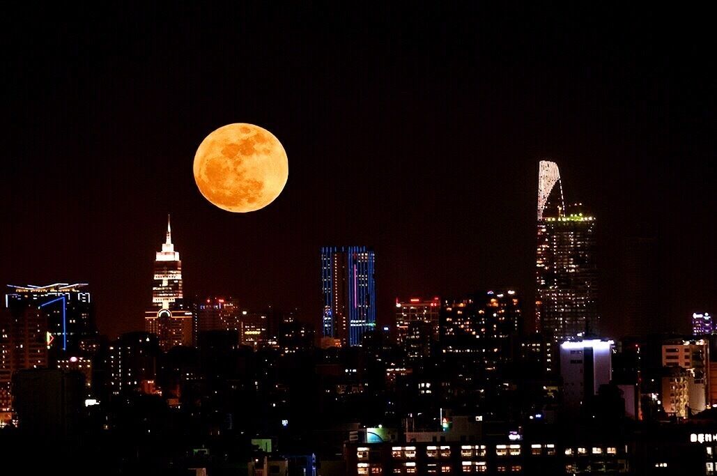 A city skyline with illuminated buildings and a full moon in the sky.