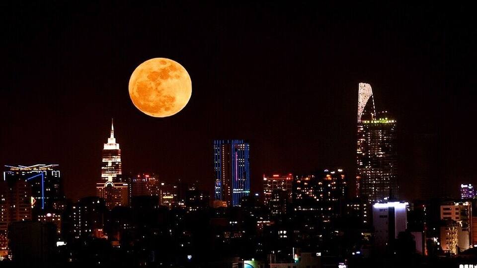 A city skyline with illuminated buildings and a full moon in the sky.
