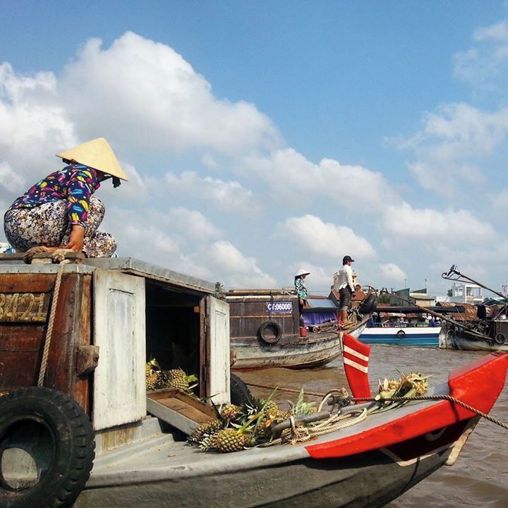 Una persona en un bote con un sombrero cónico, otra persona en un muelle, y un bote rojo y blanco en primer plano.