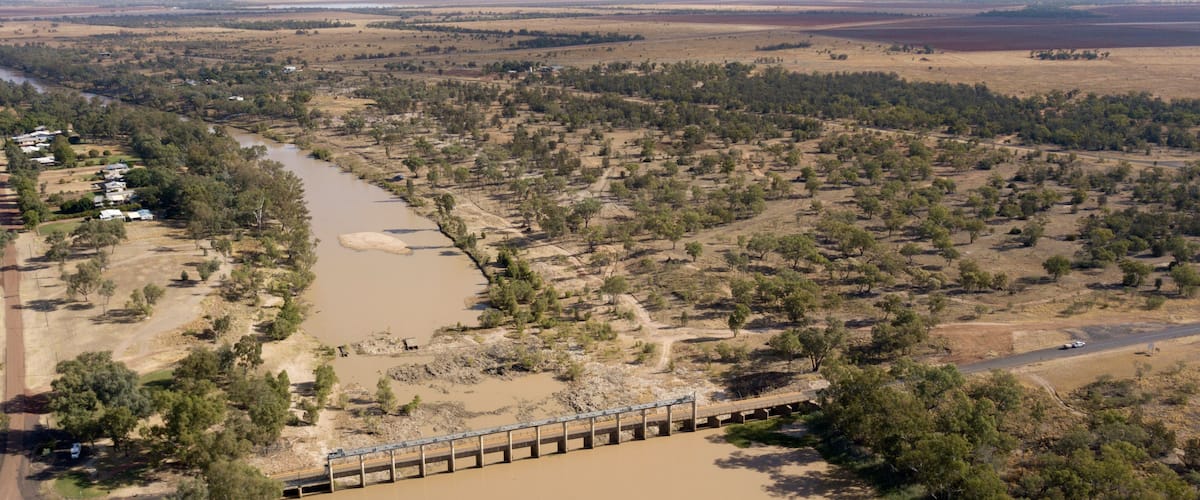 A river flowing through a dry landscape with sparse vegetation and a bridge crossing over it.