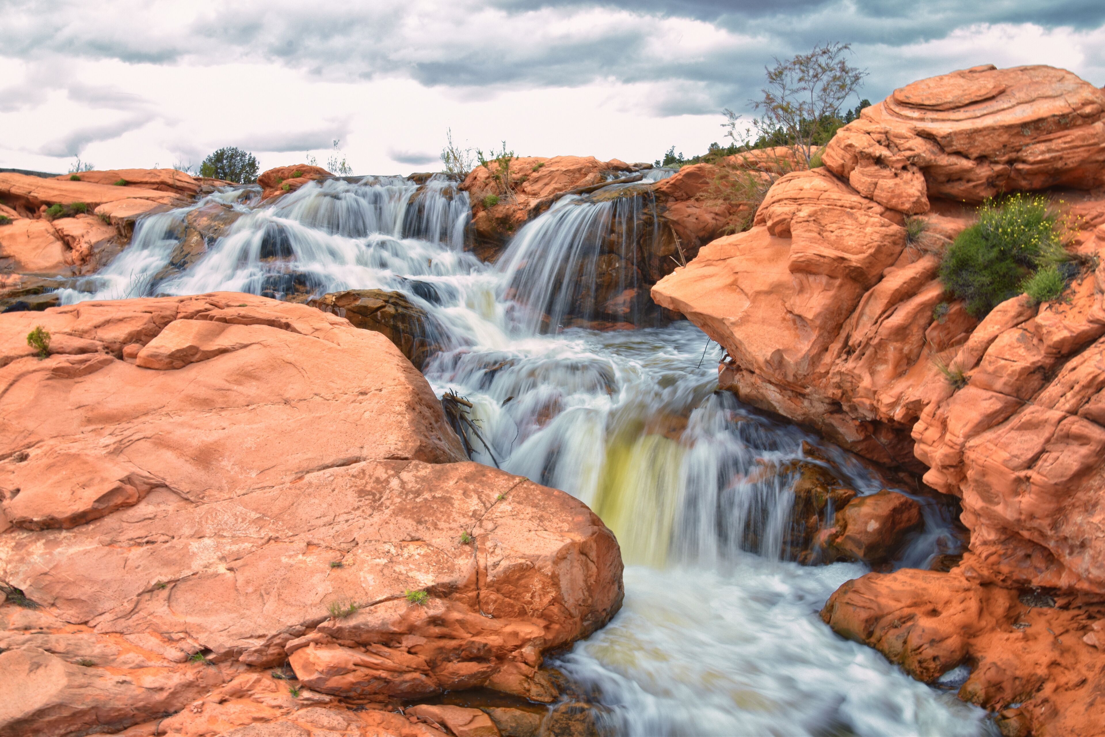 A multi-tiered waterfall cascading over reddish-brown rocks with a cloudy sky above.
