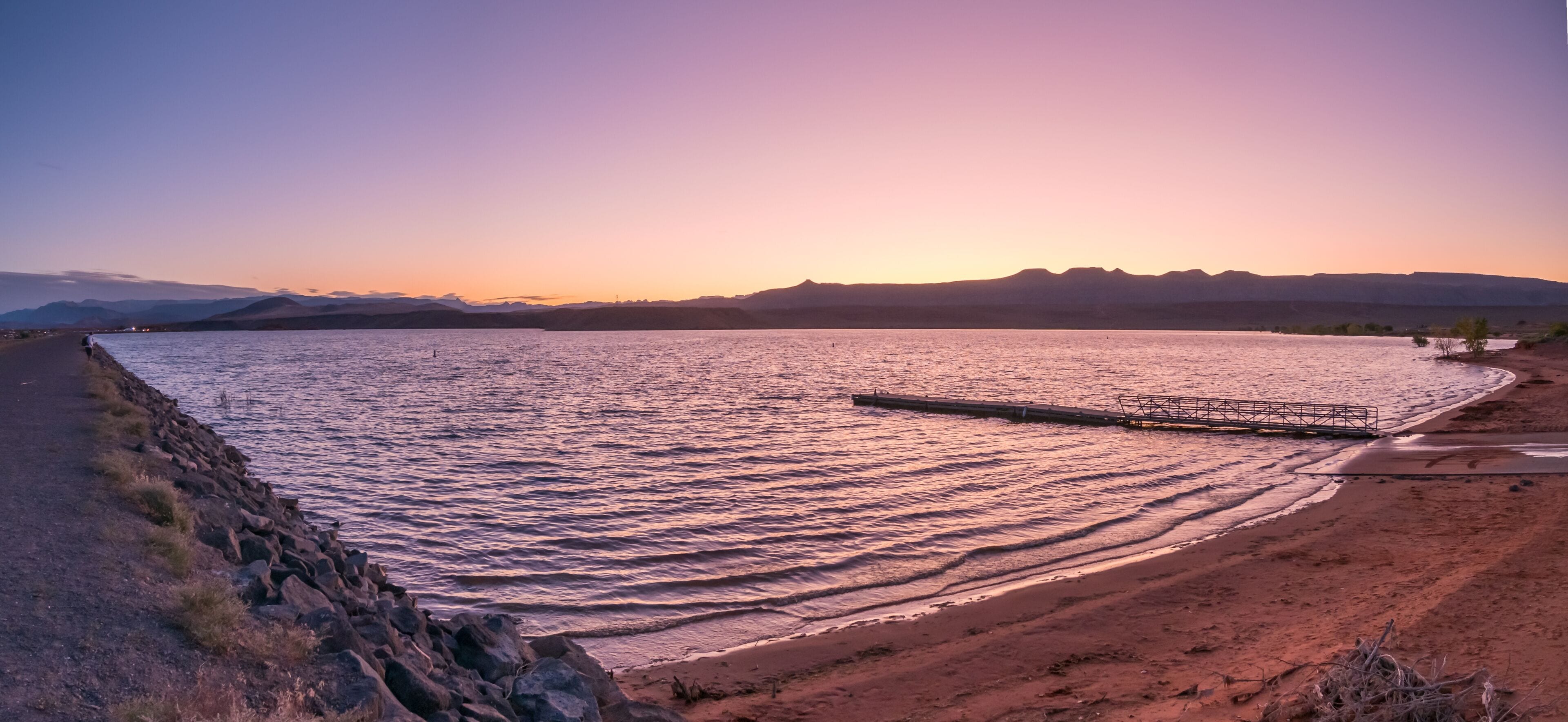 Un coucher de soleil sur un lac paisible, avec une jetée qui s'avance dans l'eau, le tout entouré de montagnes et d'une rive rocheuse.