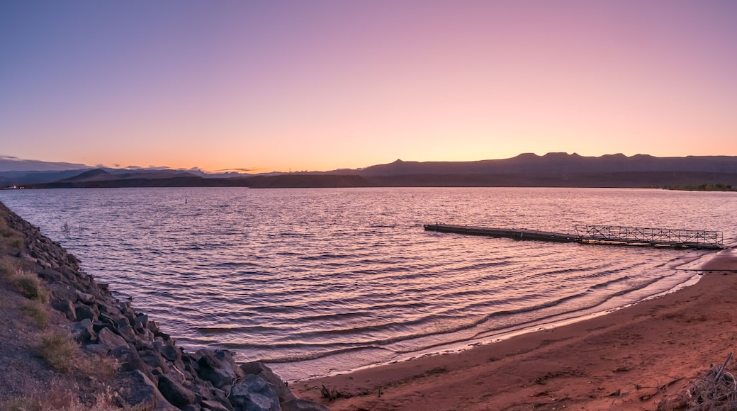 Un coucher de soleil sur un lac paisible, avec une jetée qui s'avance dans l'eau, le tout entouré de montagnes et d'une rive rocheuse.