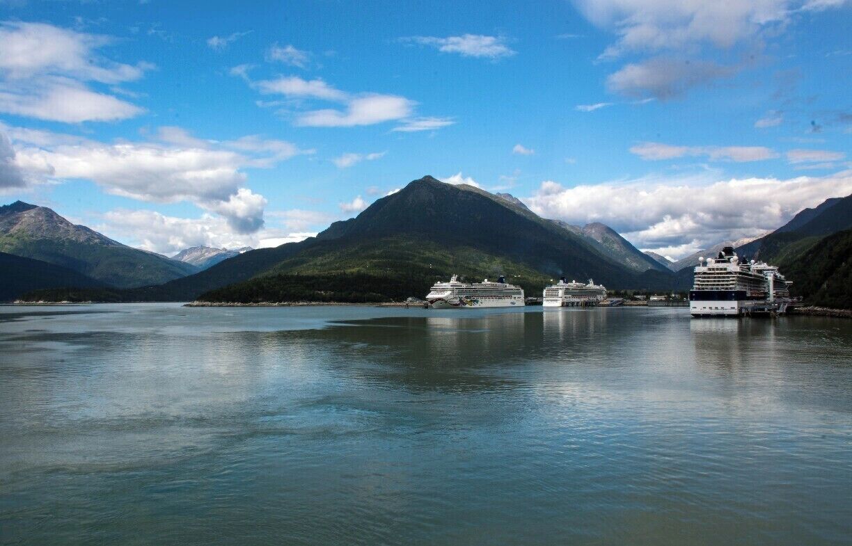 A calm fjord with two cruise ships docked, surrounded by mountains under a partly cloudy sky.