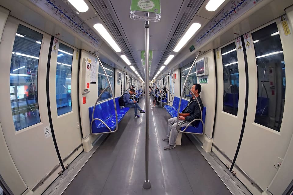 Interior de um vagão de metrô com assentos azuis, barras de apoio no teto e placas de segurança.