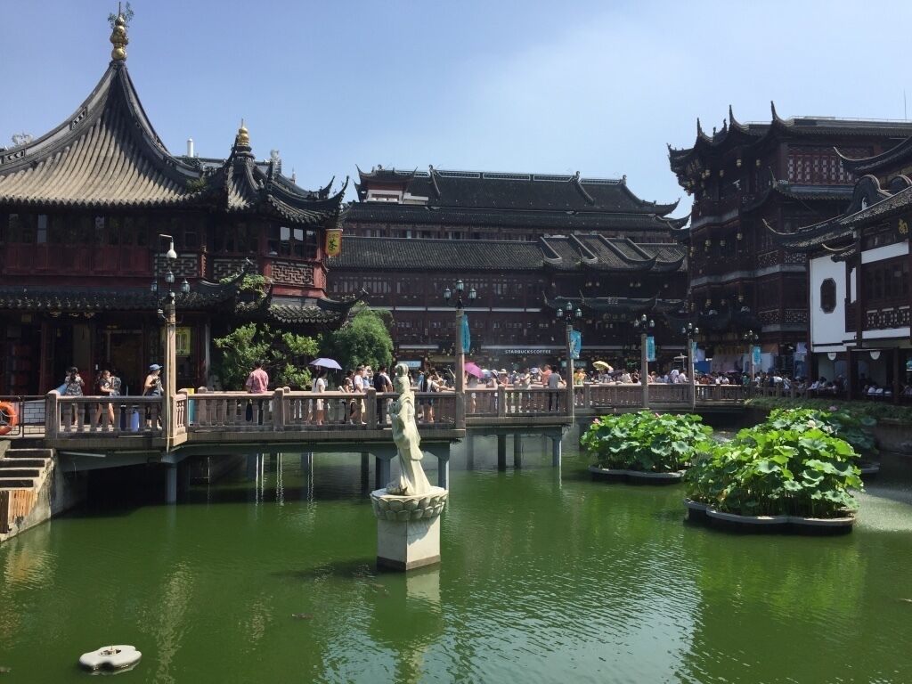 A traditional Chinese courtyard with ornate buildings, a stone bridge, and a statue in the water.