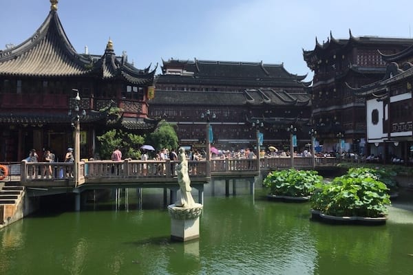 A traditional Chinese courtyard with ornate buildings, a stone bridge, and a statue in the water.