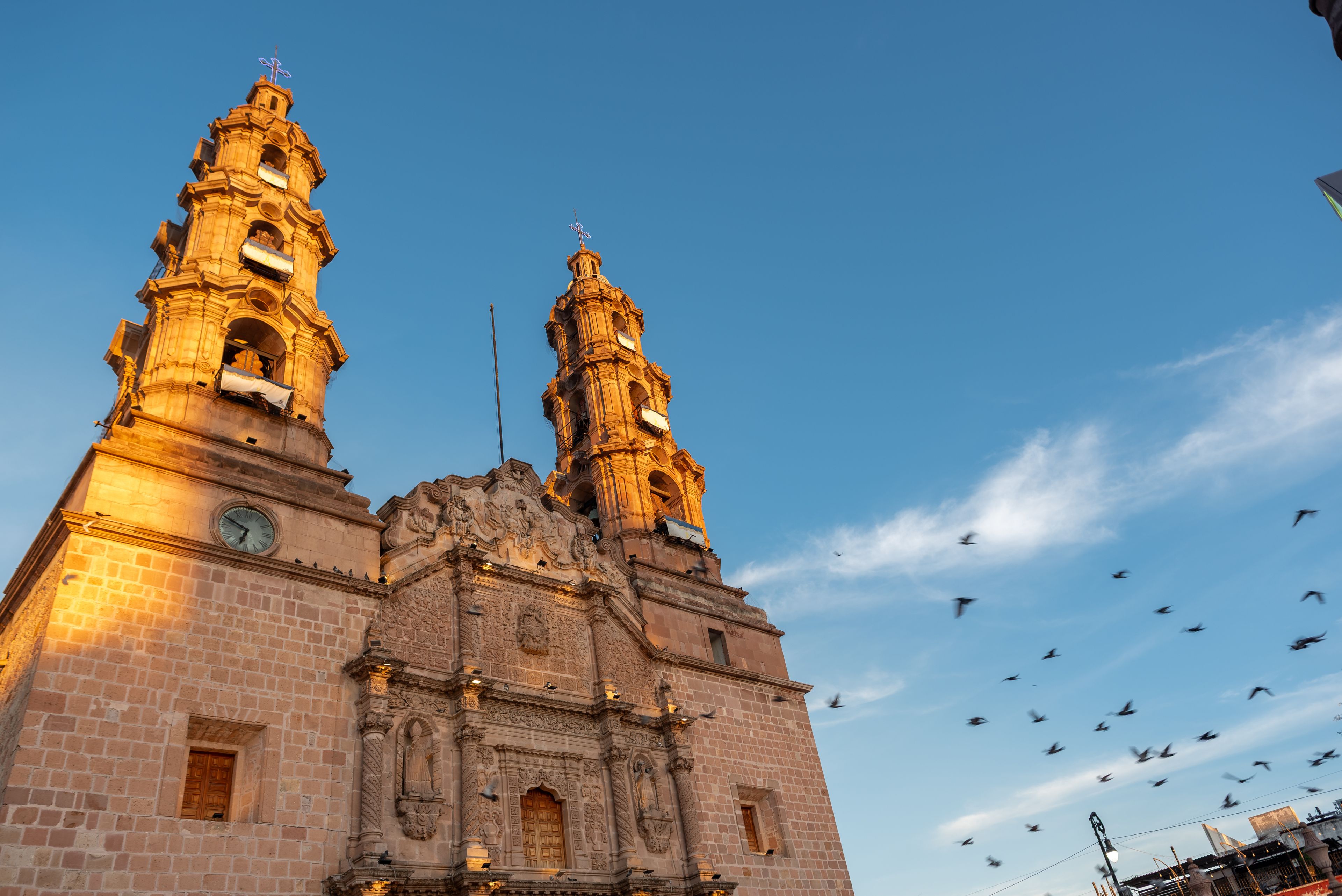 Low-angle view of the Cathedral Basilica of Our Lady of the Assumption in Aguascalientes, Mexico, at sunrise with doves flying in the sky.