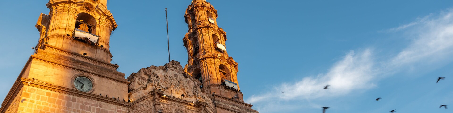 Low-angle view of the Cathedral Basilica of Our Lady of the Assumption in Aguascalientes, Mexico, at sunrise with doves flying in the sky.