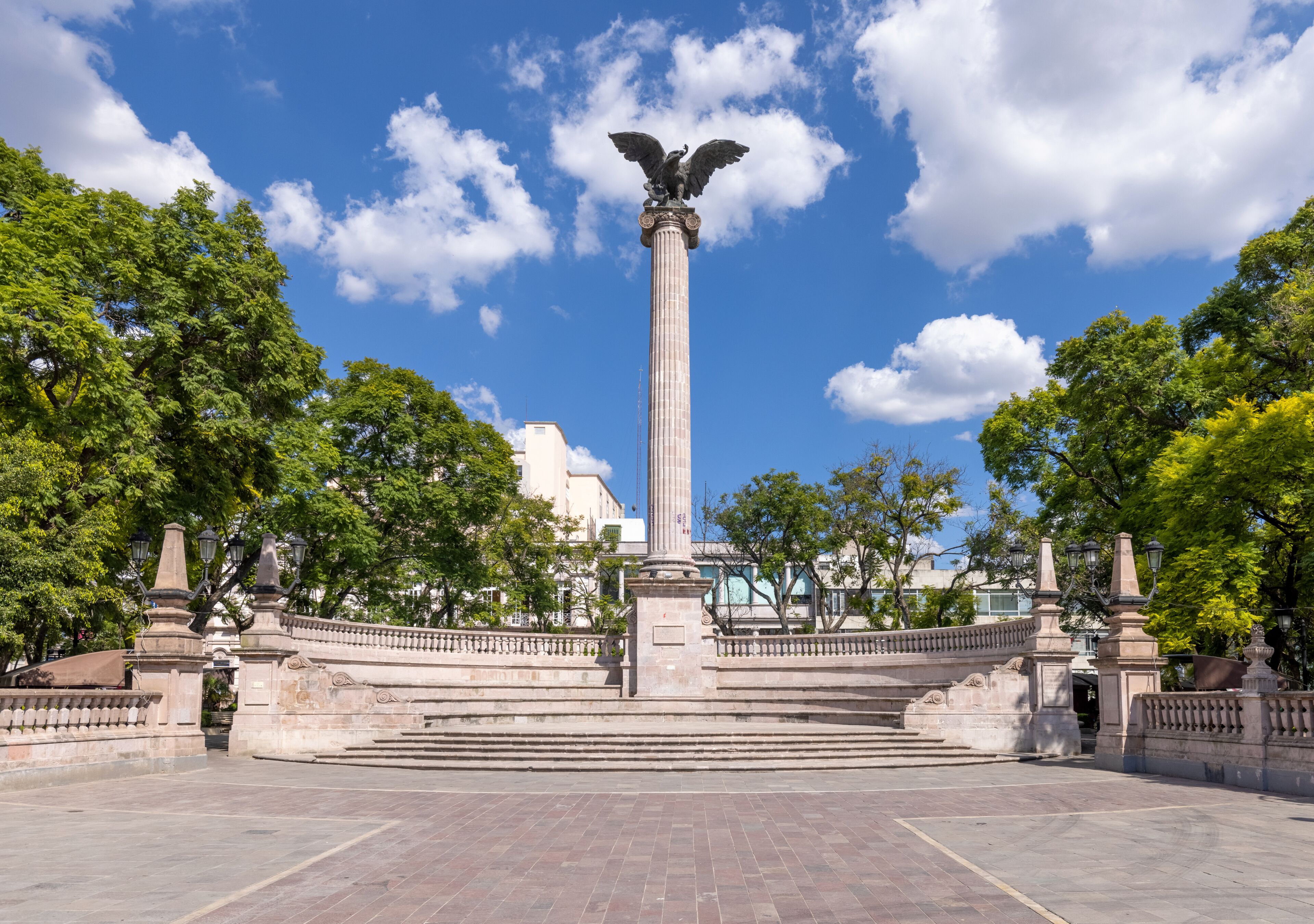 A tall monument with an eagle statue, surrounded by a circular plaza with steps leading up to it, and trees in the background.