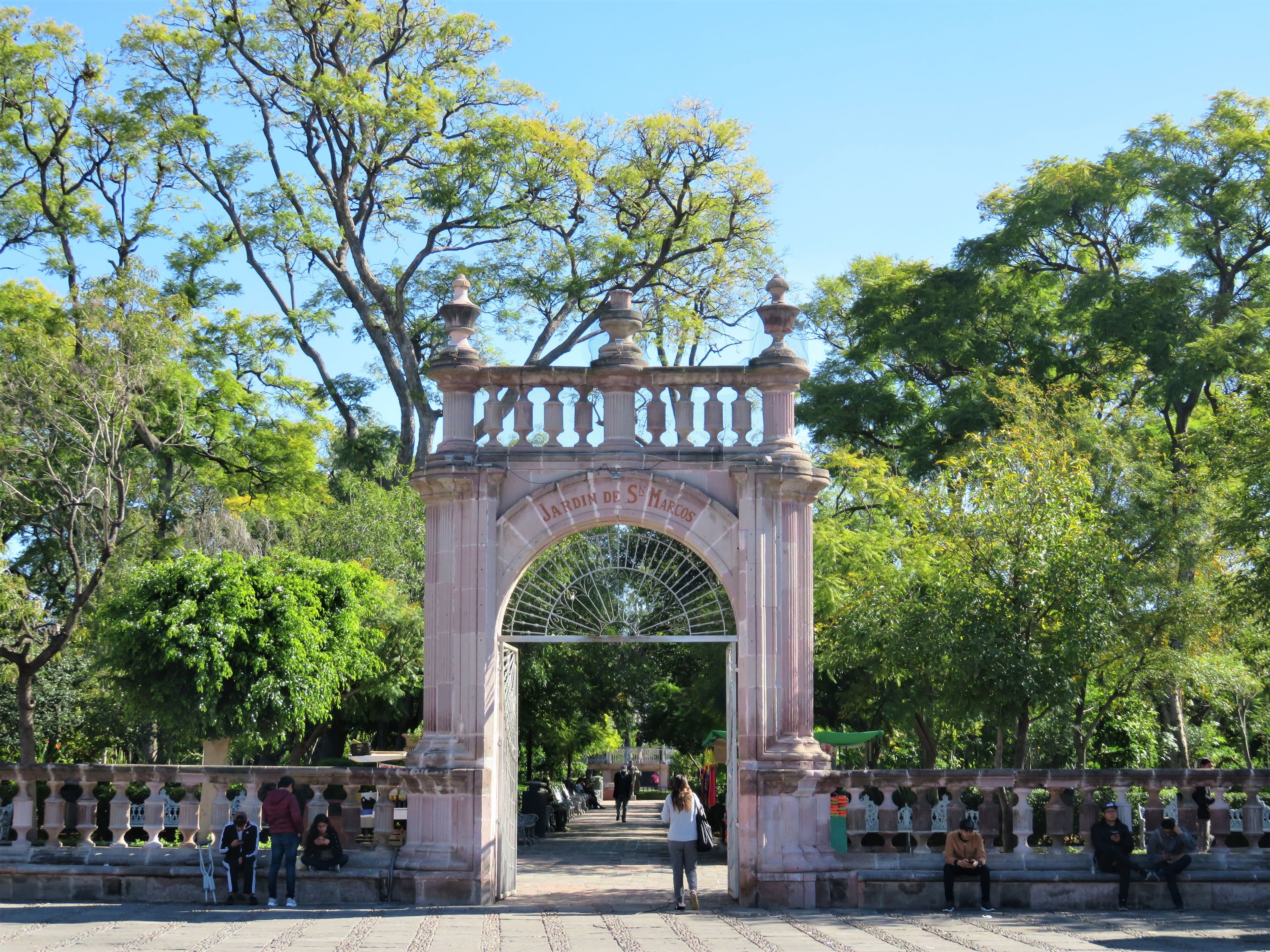 Arcada de pedra ornamentada com a inscrição 'JARDIN DE SE MARCOS' dando acesso a um jardim exuberante.