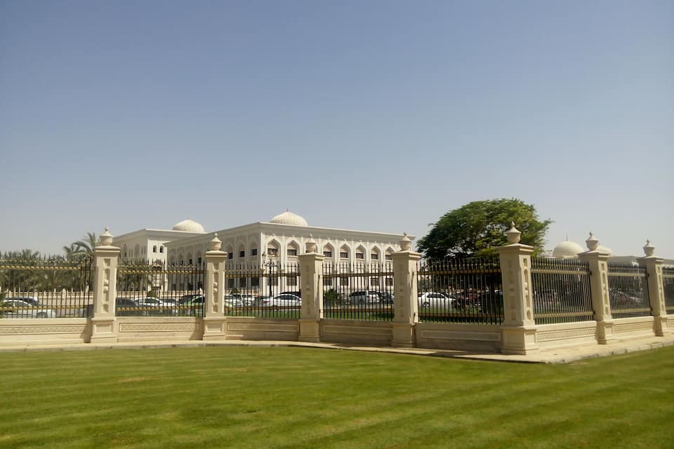 A large, white building with domes, surrounded by a fence and palm trees.