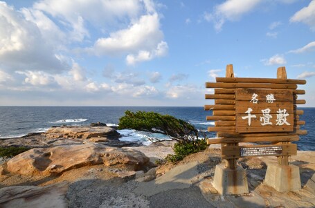 Ein Holzschild mit chinesischen Schriftzeichen, eine felsige Küstenlinie und das Meer im Hintergrund.