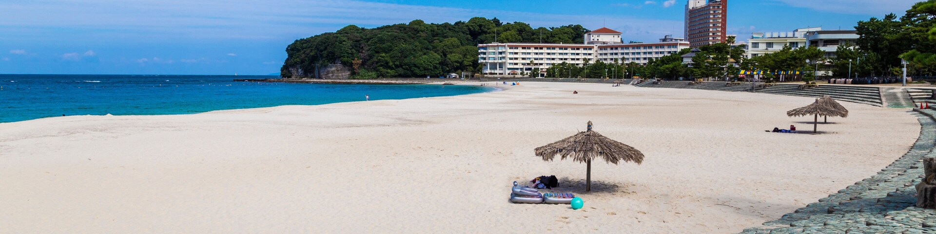 Una playa con agua azul cristalina, una orilla arenosa, un paraguas de paja y un camino de piedra.