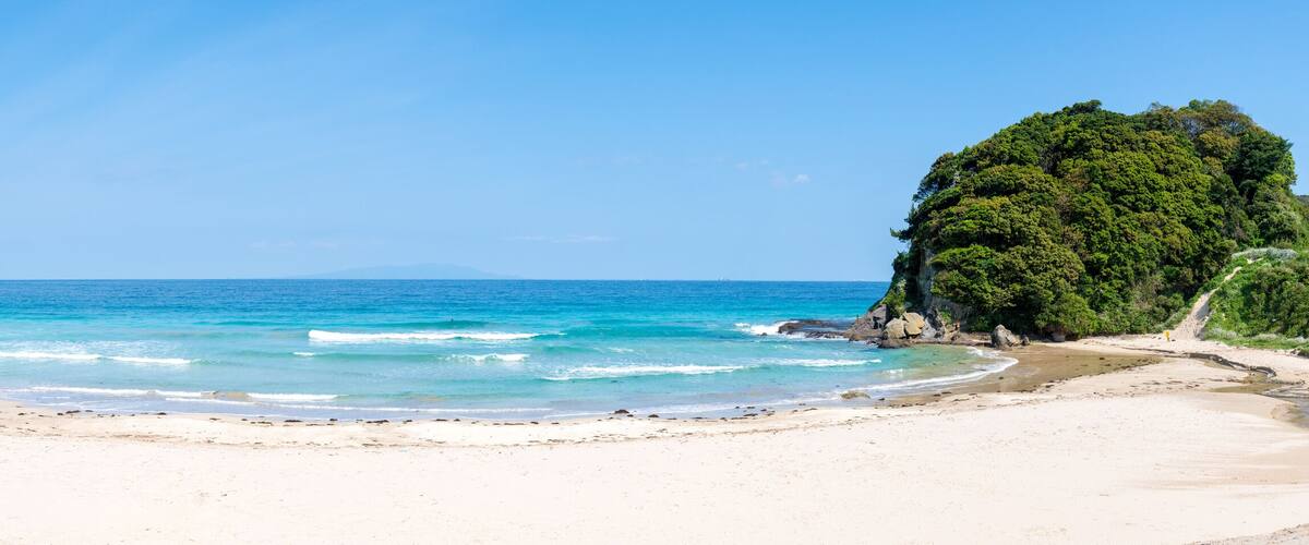 A beach with clear blue waters, sandy shore, and a large green hill.