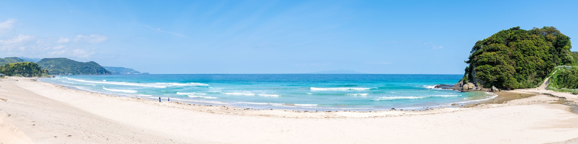 A beach with clear blue waters, sandy shore, and a large green hill.