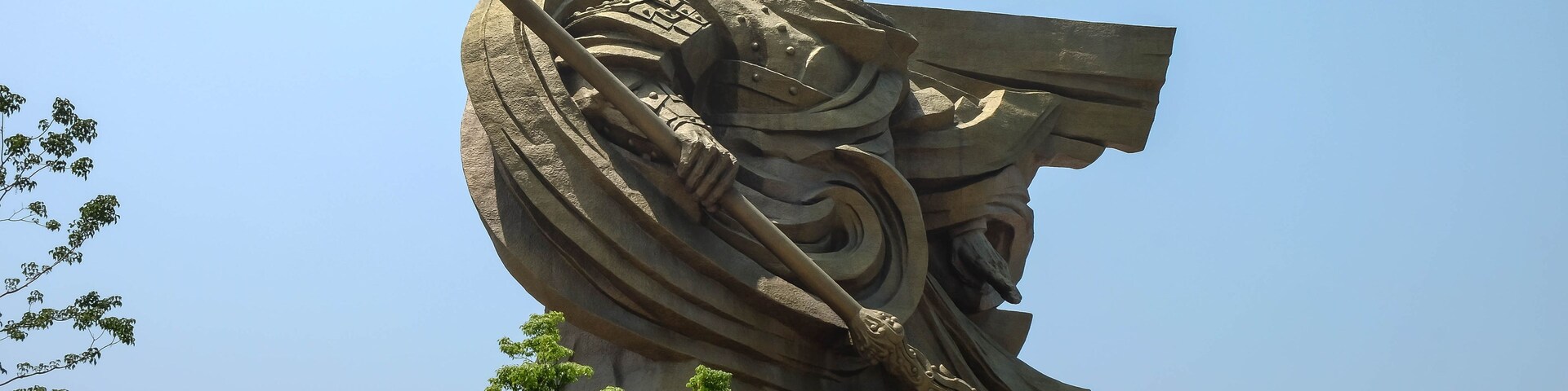 Estátua grande e elaborada de um guerreiro com uma lança, ao fundo um céu azul límpido.