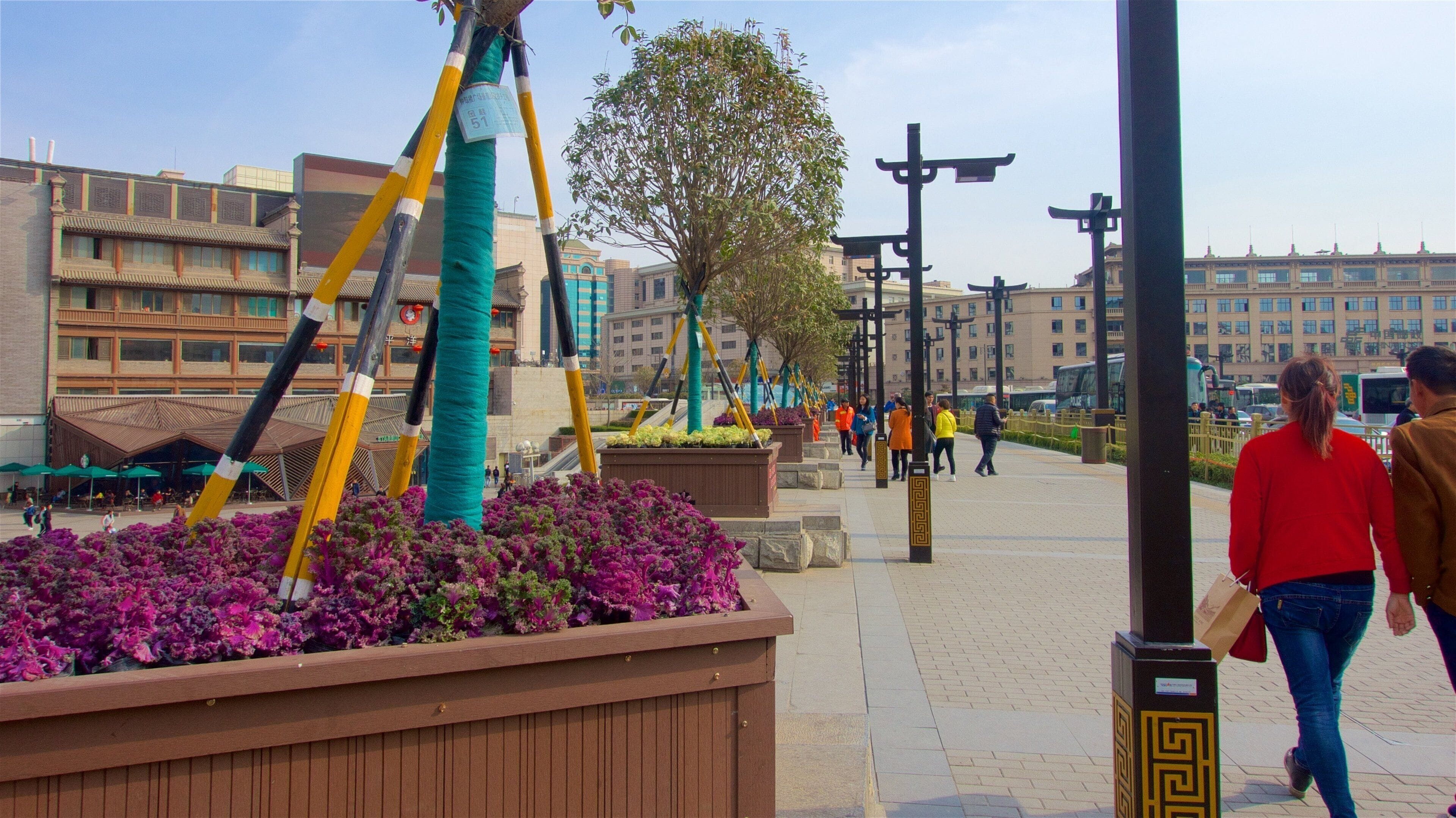 Una plaza pública con arquitectura moderna y tradicional, incluyendo un edificio de techo tradicional y fachada de vidrio moderna. Se ven personas caminando, una fila de farolas y un árbol en el centro.