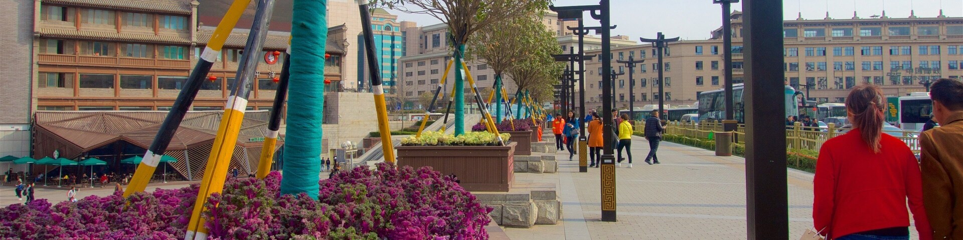 Una plaza pública con arquitectura moderna y tradicional, incluyendo un edificio de techo tradicional y fachada de vidrio moderna. Se ven personas caminando, una fila de farolas y un árbol en el centro.