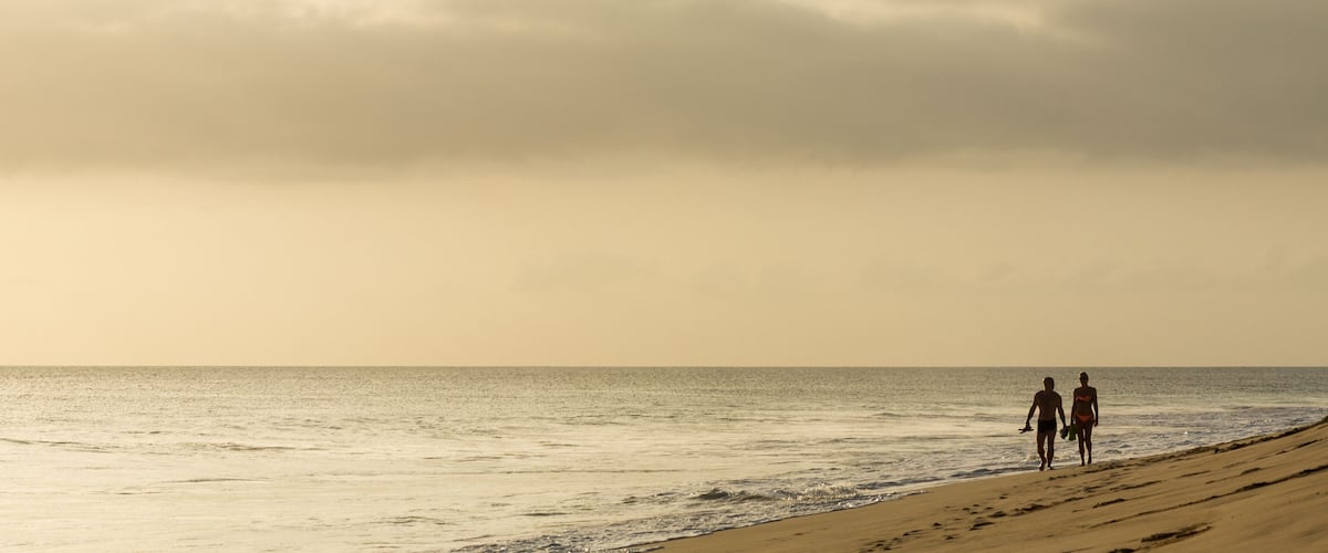 A beach with two people walking along the shore, waves crashing onto the sand, and a cloudy sky.