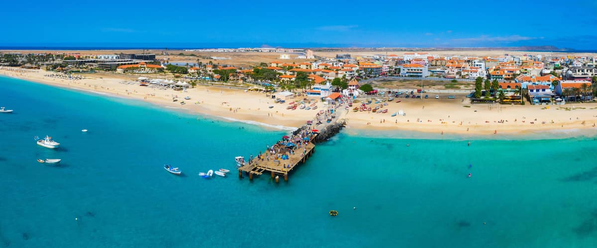 A coastal town with a pier extending into turquoise waters, a sandy beach, and a backdrop of buildings.