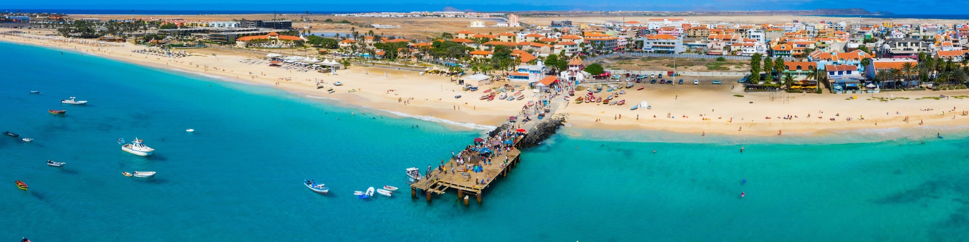 A coastal town with a pier extending into turquoise waters, a sandy beach, and a backdrop of buildings.