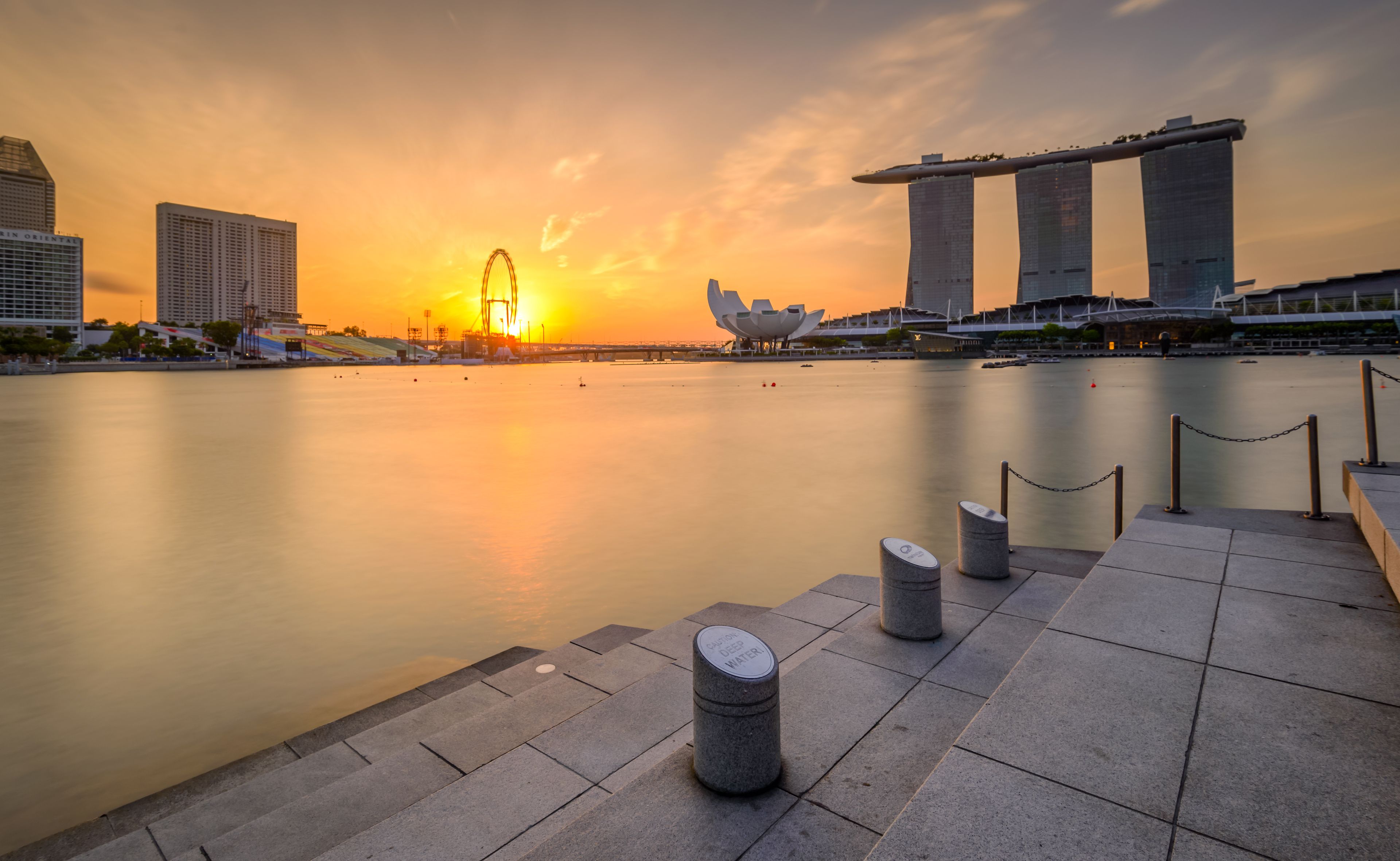 Un paisaje urbano frente al agua al atardecer, con edificios destacados, incluyendo una estructura moderna distintiva y el hotel Marina Bay Sands.