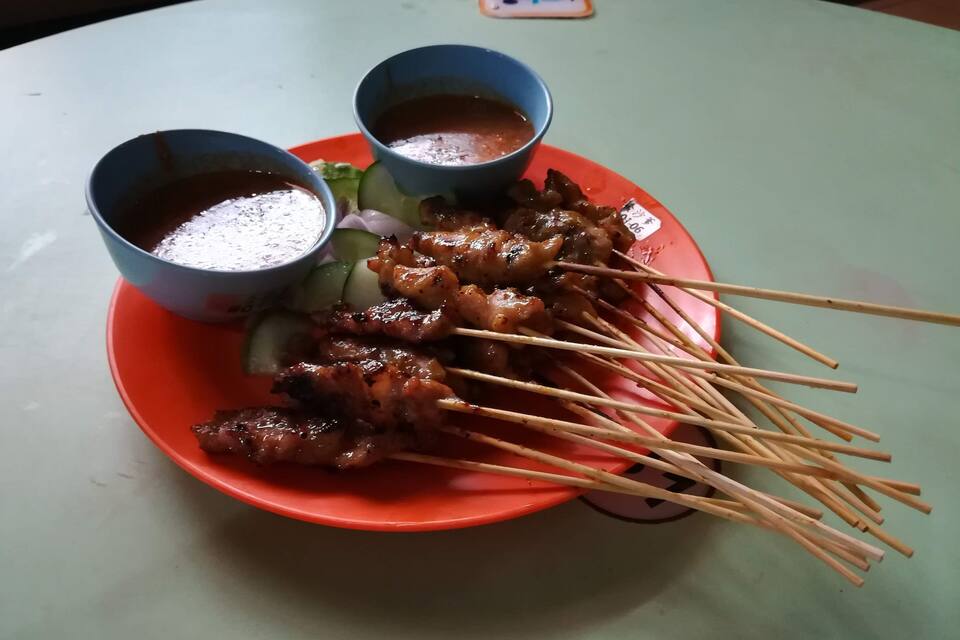 A plate of grilled meat skewers with dipping sauces and cucumber slices on a table.