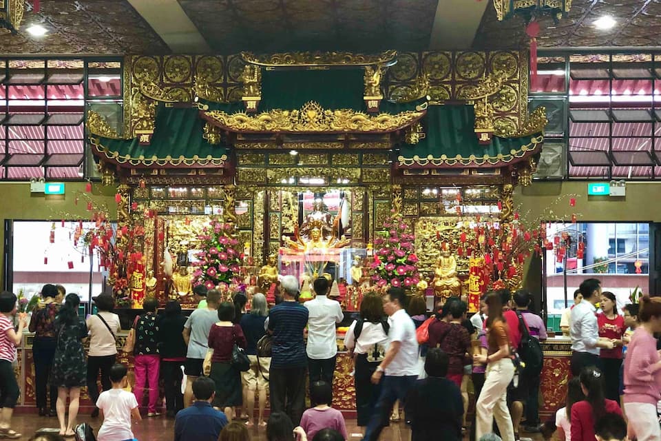 A crowded temple with ornate golden decorations and floral arrangements.