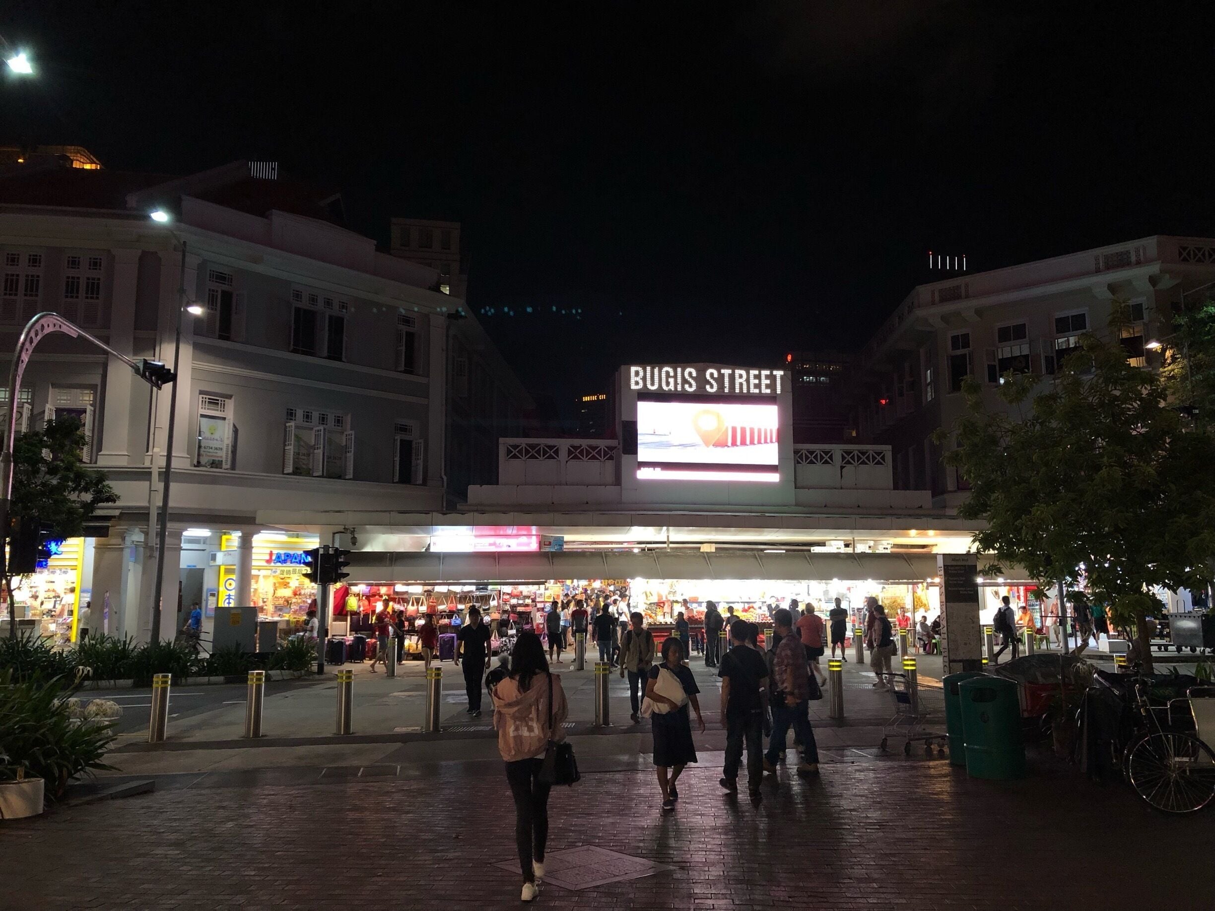 Une rue animée de nuit, avec une enseigne bien visible indiquant « BUGIS STREET » et des devantures de magasins illuminées.