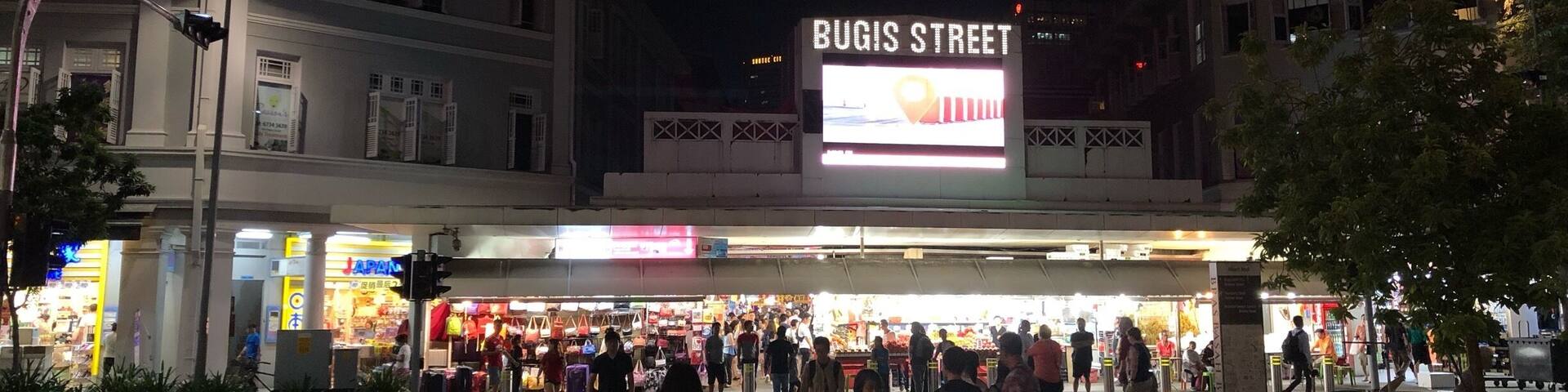 A busy street scene at night with a prominent 'BUGIS STREET' sign and illuminated storefronts.