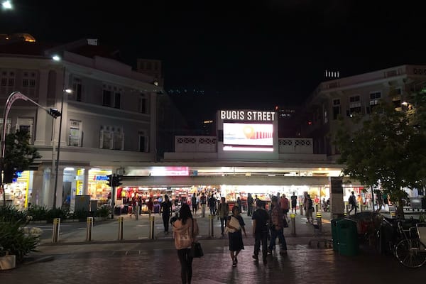 A busy street scene at night with a prominent 'BUGIS STREET' sign and illuminated storefronts.