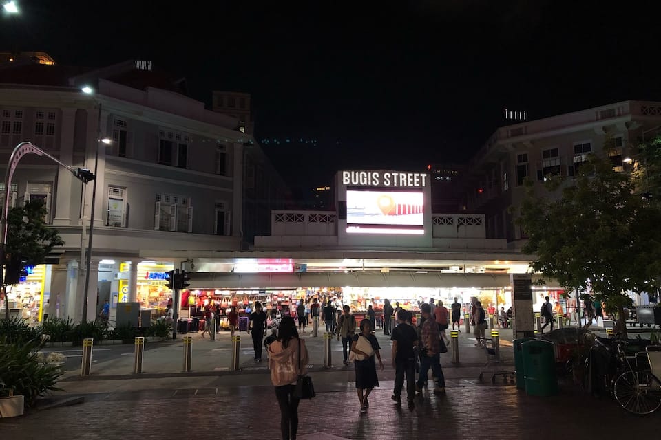Cena movimentada de rua à noite, com uma placa grande com a inscrição 'BUGIS STREET' e vitrines iluminadas.