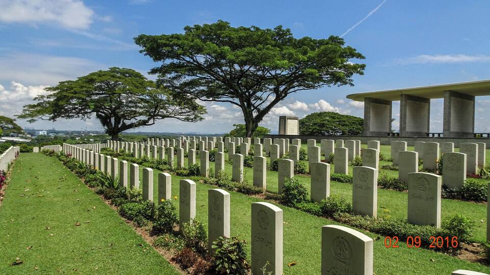 Ein Friedhof mit Reihen von Grabsteinen, einem großen Baum und einem Gebäude im Hintergrund.