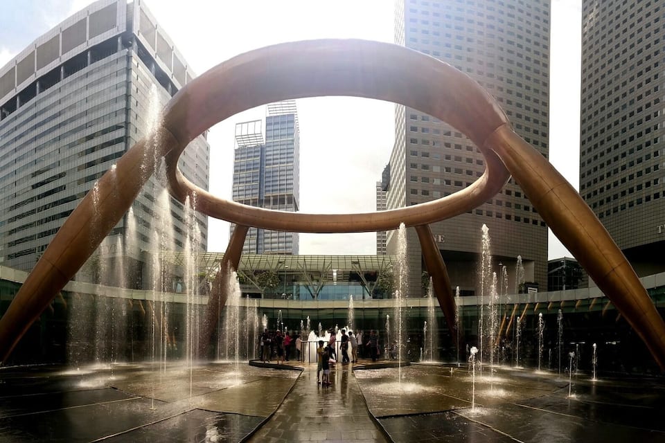 Una plaza urbana moderna con una gran fuente circular y edificios altos al fondo.