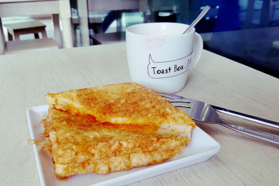 Sándwich de queso a la plancha sobre un plato blanco, con una taza tipo caja de tostadas al fondo.