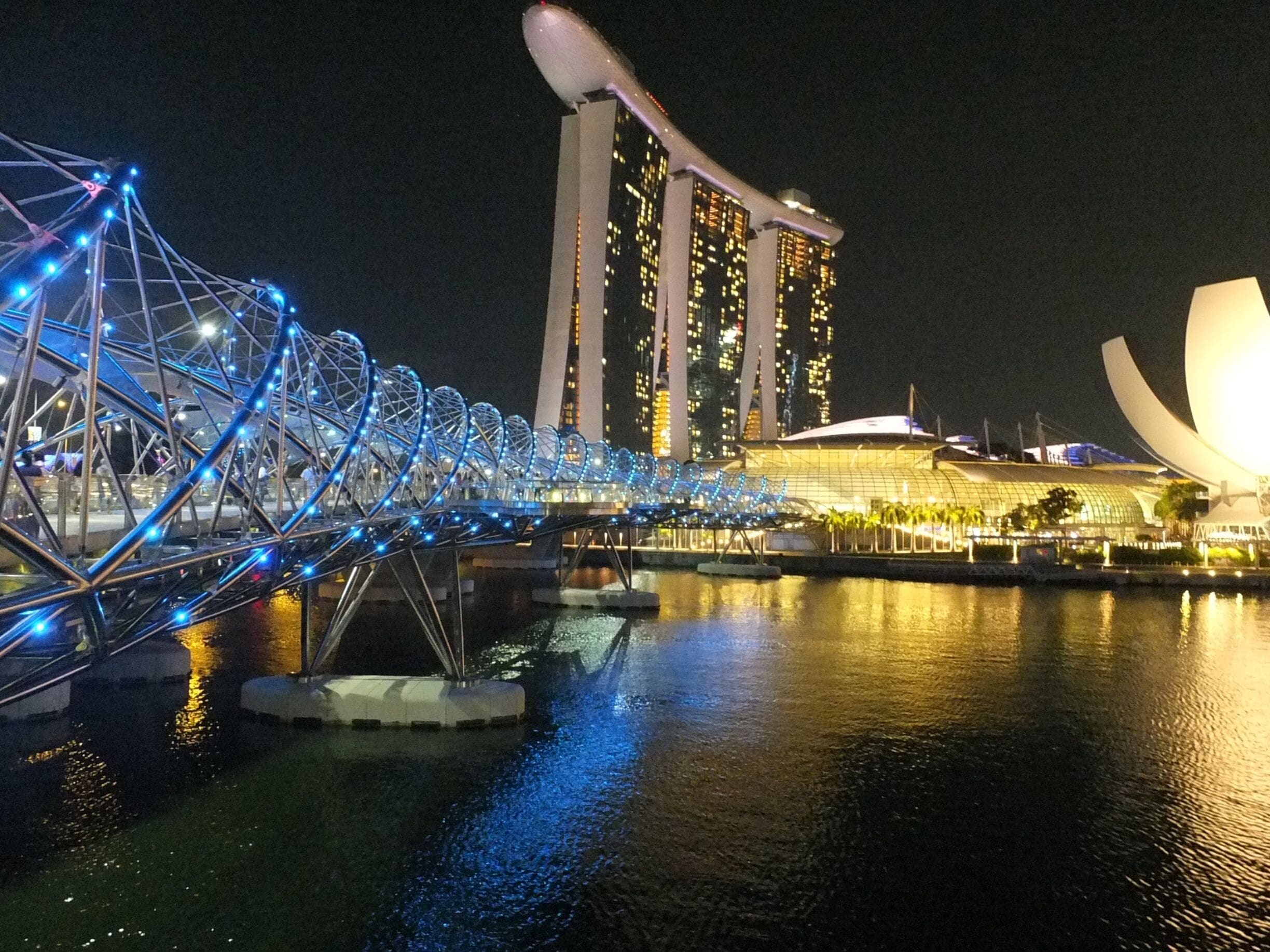 Vista nocturna de un puente con luces azules, un edificio moderno con numerosas ventanas y un edificio de forma curva y fachada blanca.