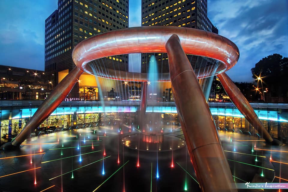 Una escultura moderna con base circular y tres pilares altos, rodeada por una plaza con luces láser de colores.