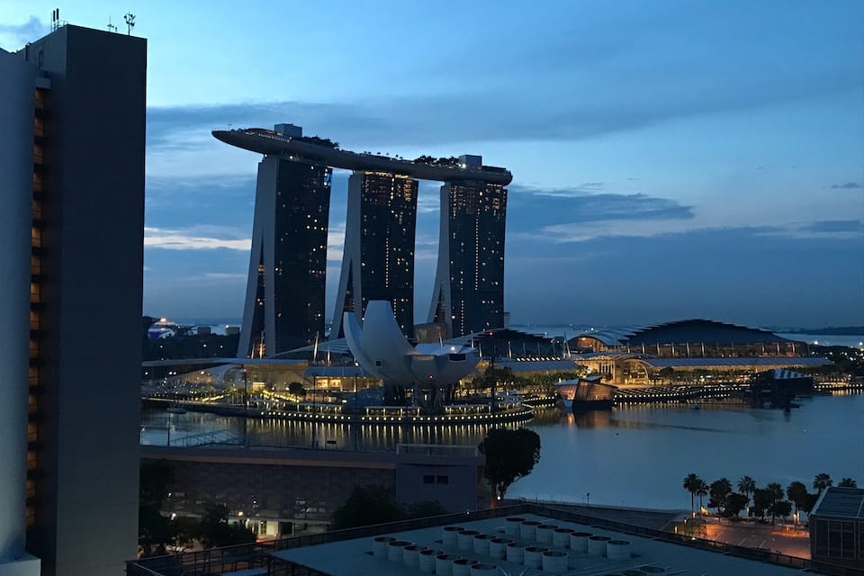 A cityscape with a prominent modern skyscraper and a unique sculpture near a waterfront.