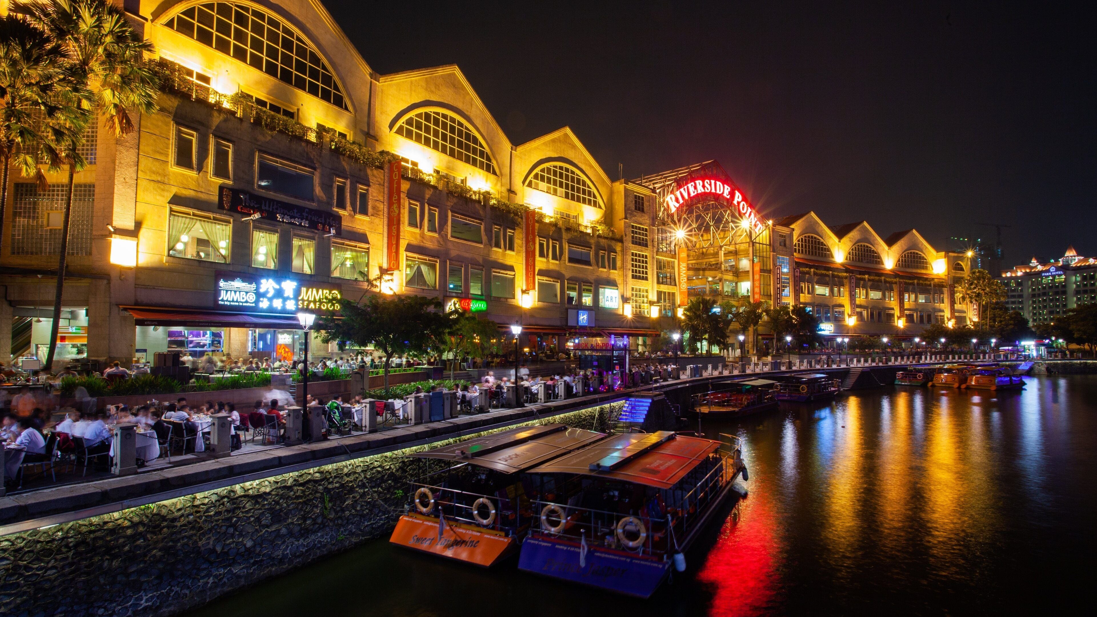 A riverside promenade at night with illuminated buildings, a bridge, and boats.