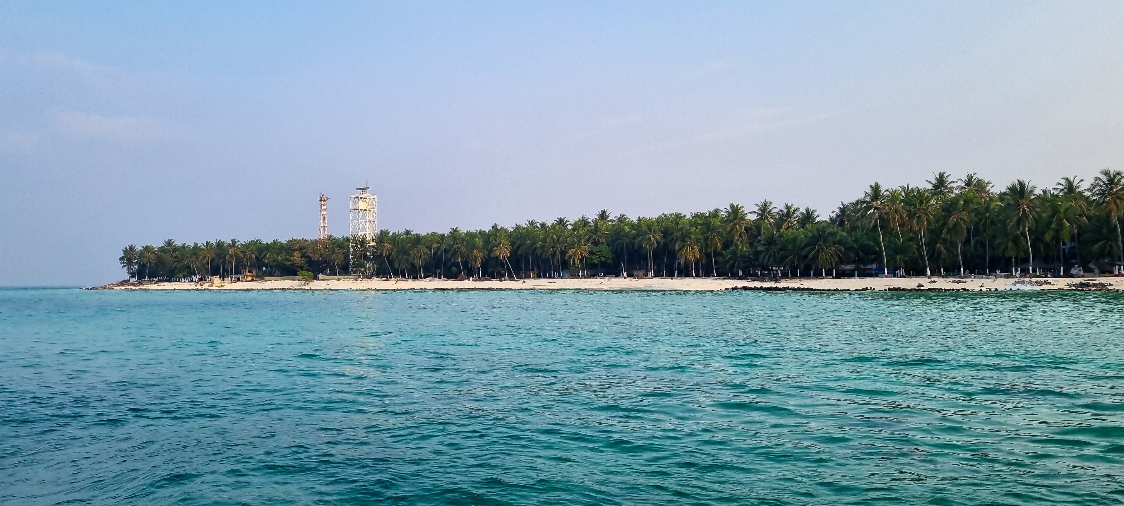 A coastal landscape with a sandy beach, a line of palm trees, and a tall tower structure.