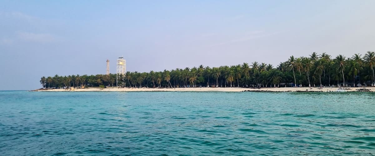 A coastal landscape with a sandy beach, a line of palm trees, and a tall tower structure.