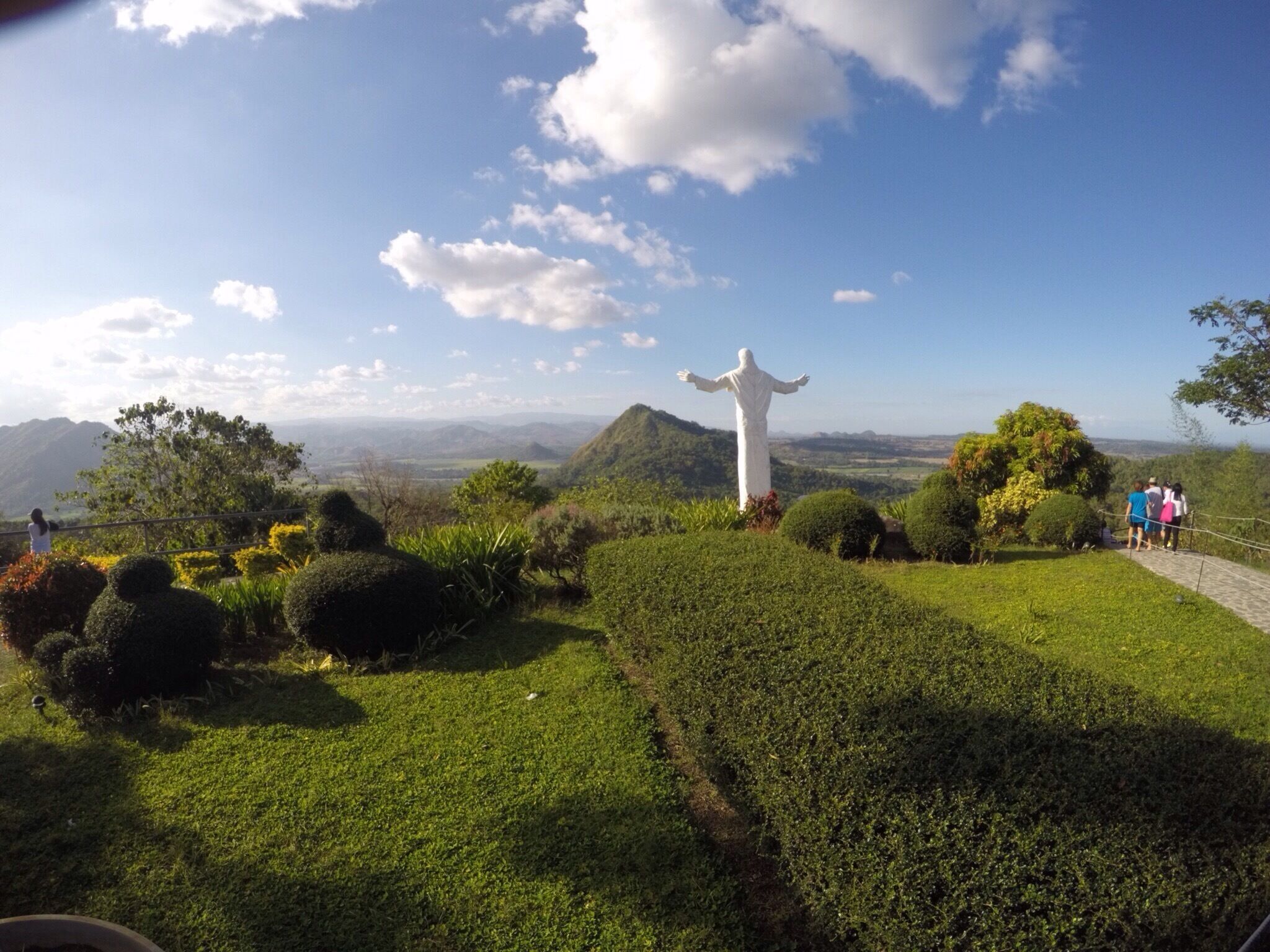 A statue of a figure with outstretched arms on a grassy hill with a scenic view of mountains and a clear sky.