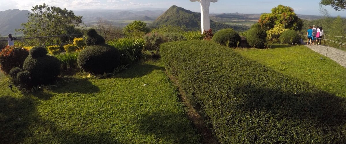 A statue of a figure with outstretched arms on a grassy hill with a scenic view of mountains and a clear sky.