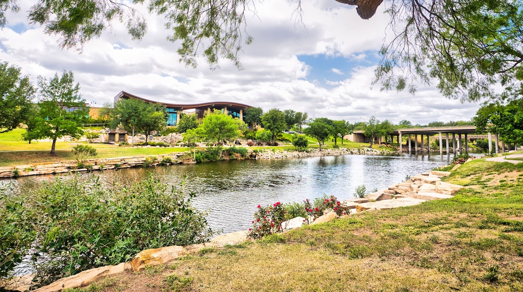 A visitor center on the other side of a tranquil river in San Angelo, Texas, surrounded by lush grass and trees with wispy clouds dotting a bright blue sky.