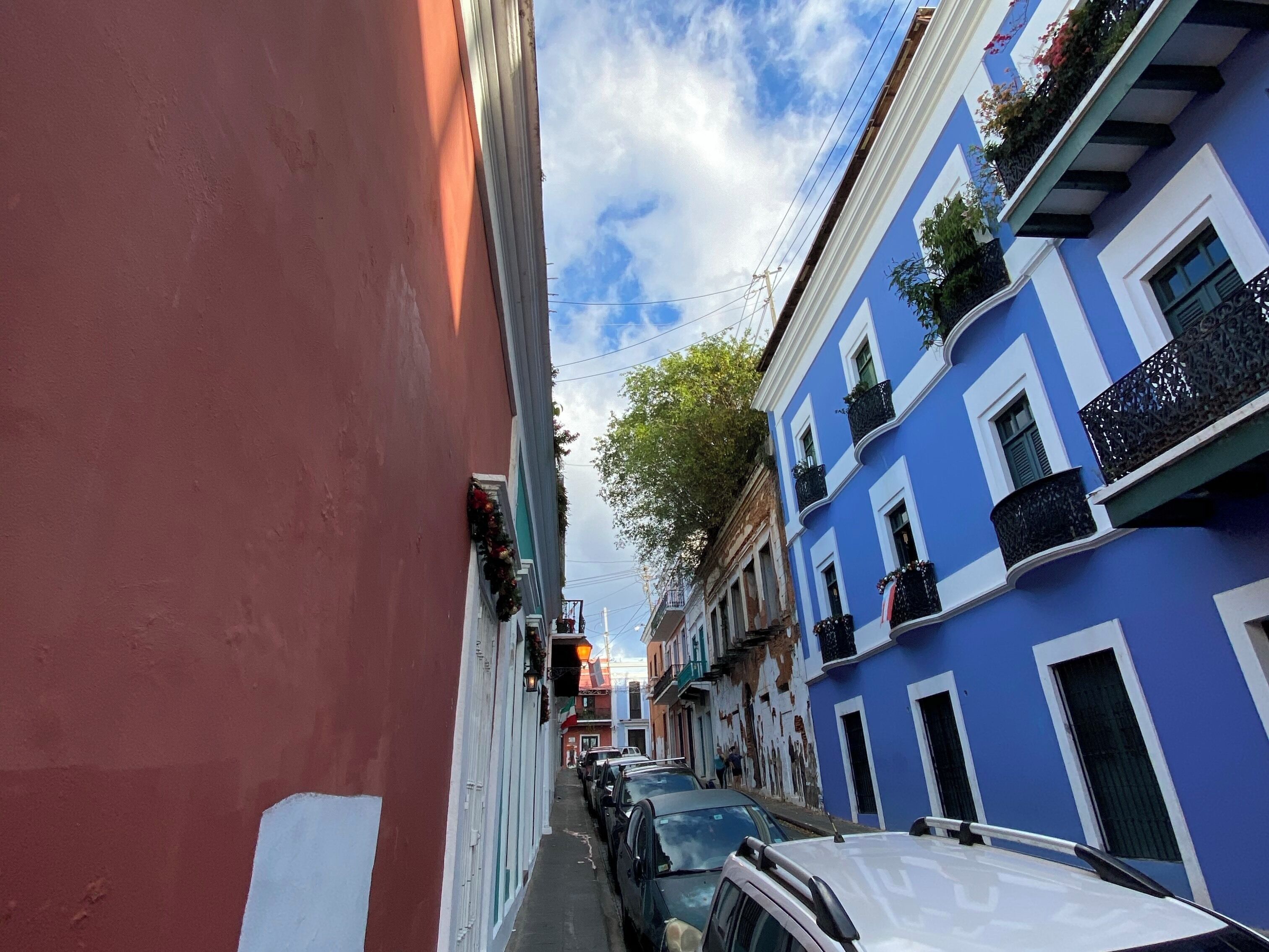 A narrow street with colorful buildings, parked cars, and a clear sky.