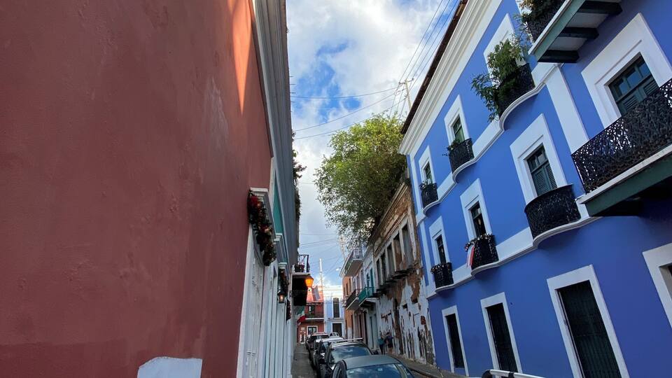 A narrow street with colorful buildings, parked cars, and a clear sky.