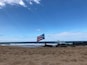 A beach with a flag on a post, waves crashing, and a clear sky.