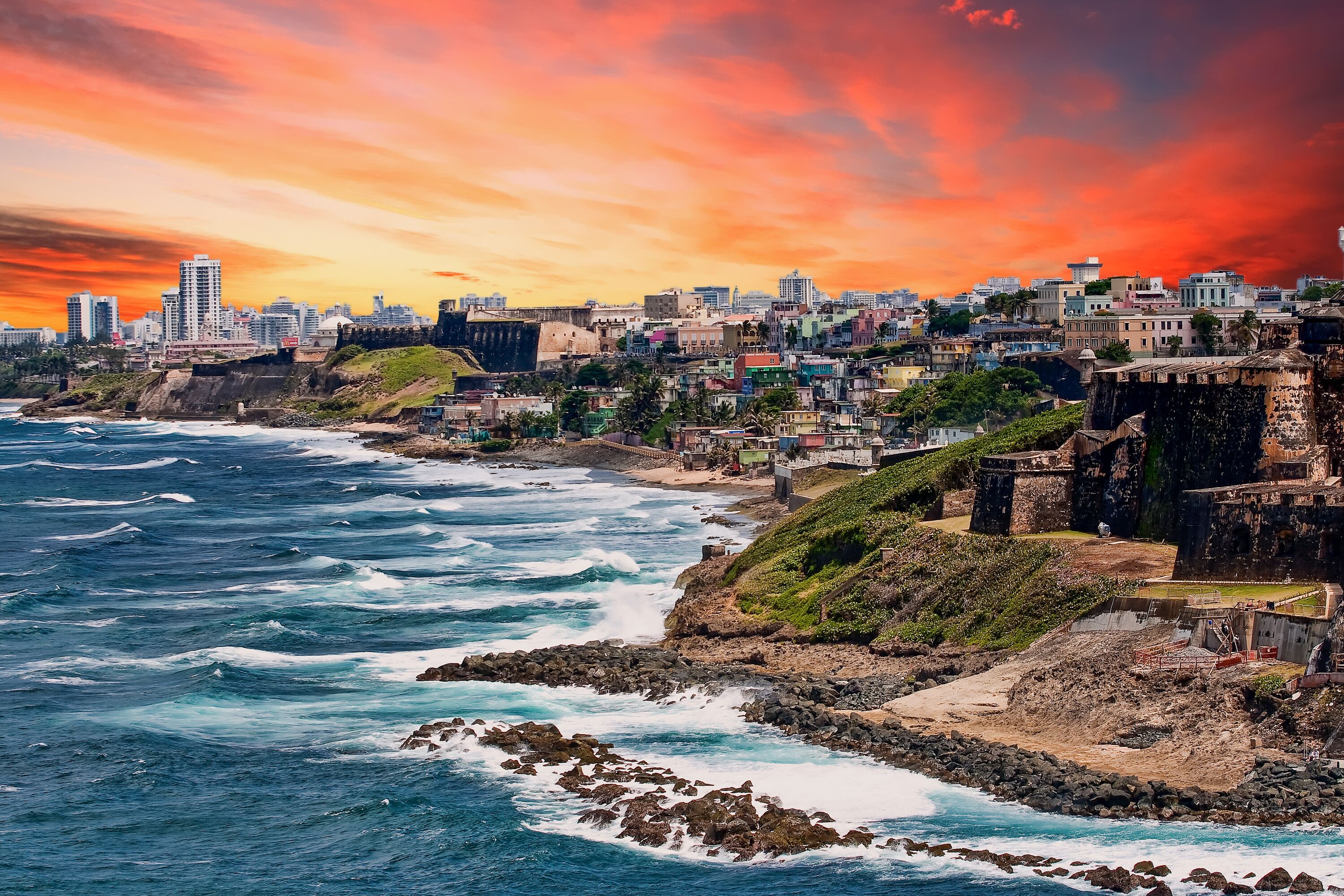 A coastal cityscape with waves crashing against rocky shores, historic buildings, and a sunset sky.